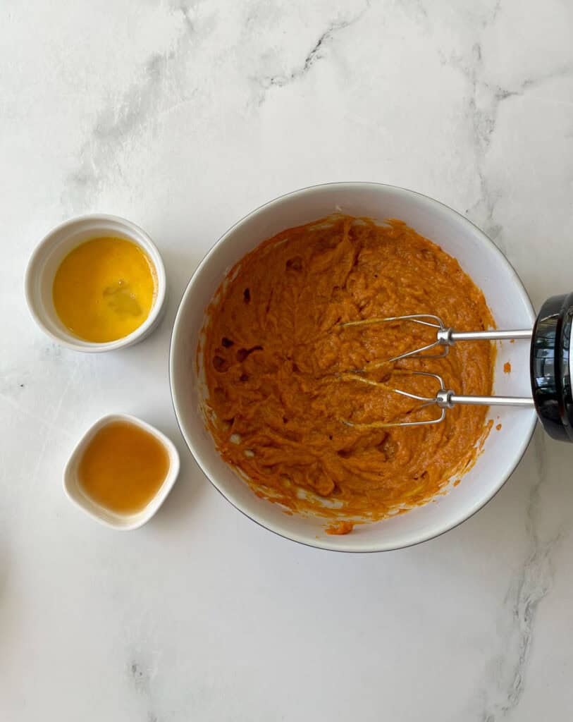 A bowl of creamy pumpkin puree mixture, accompanied by small bowls of slightly beaten egg and vanilla.