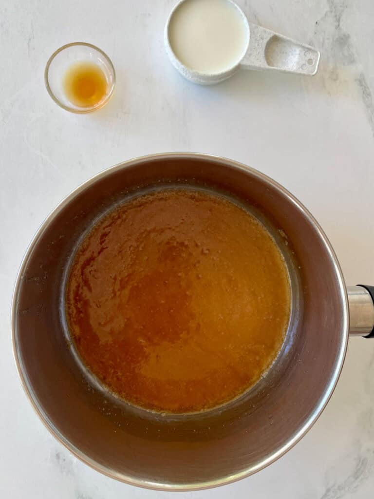 Caramel sauce cooking in a saucepan with milk and vanilla extract nearby.