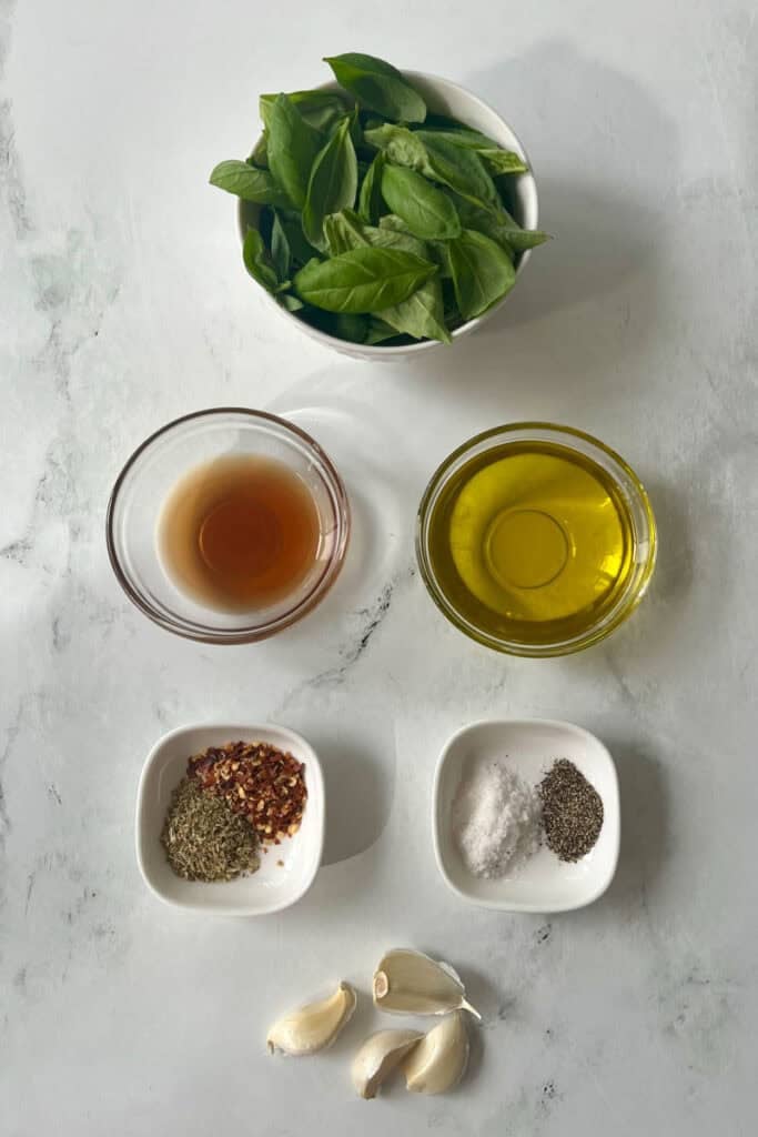 Fresh basil leaves with garlic, olive oil, and seasonings for chimichurri sauce on a white surface.