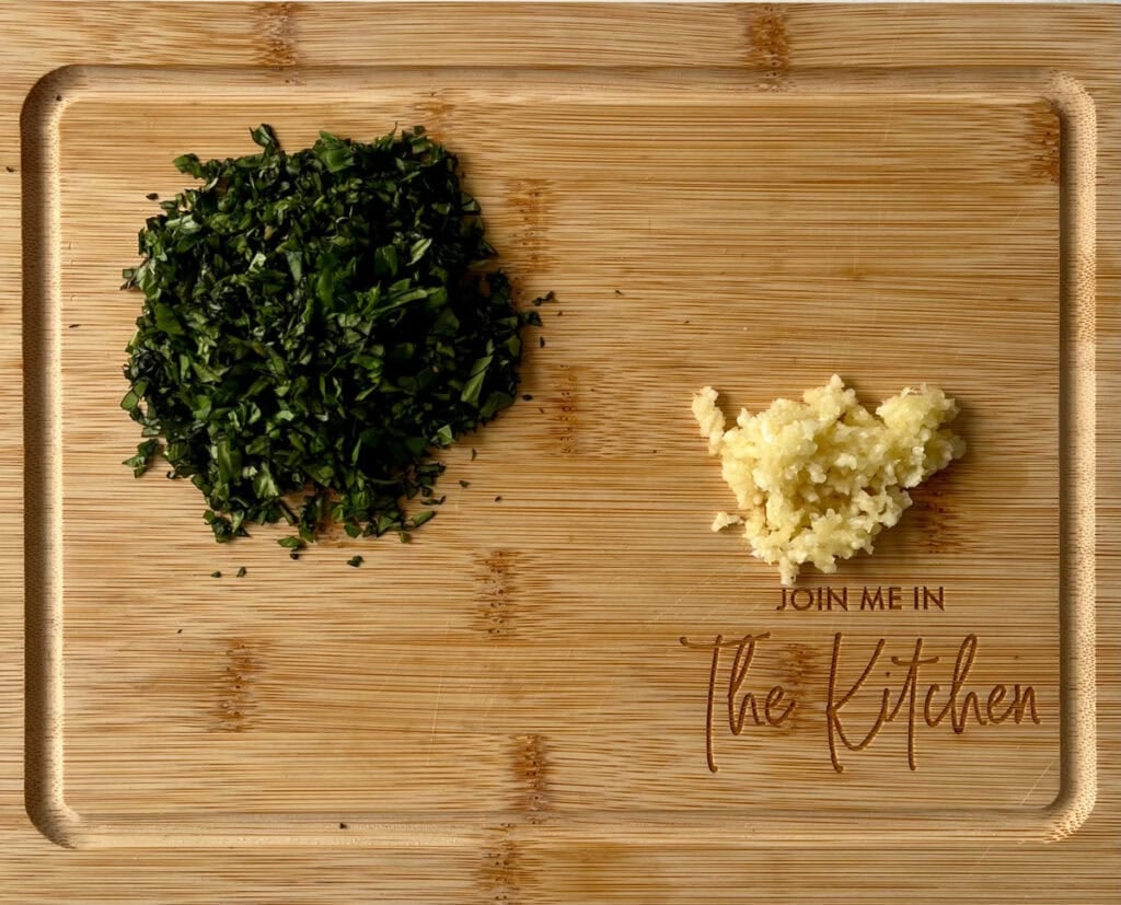 Chopped herbs and minced garlic on wooden cutting board for cooking.