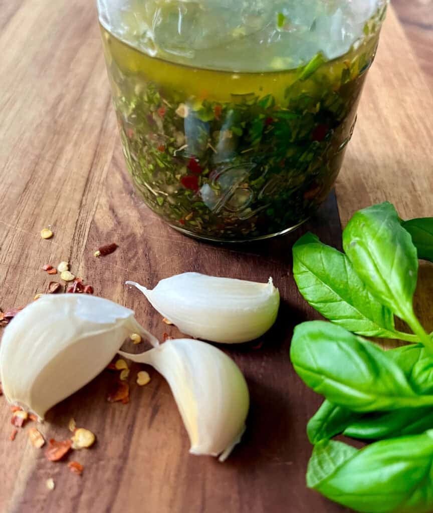 Fresh basil, garlic, and homemade chimichurri sauce in a glass jar on wooden surface.