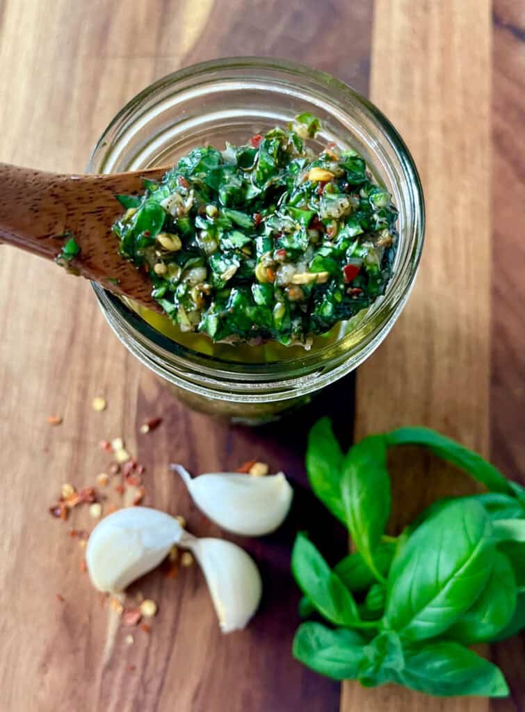 Fresh homemade basil chimichurri sauce in a glass jar with garlic and basil nearby.