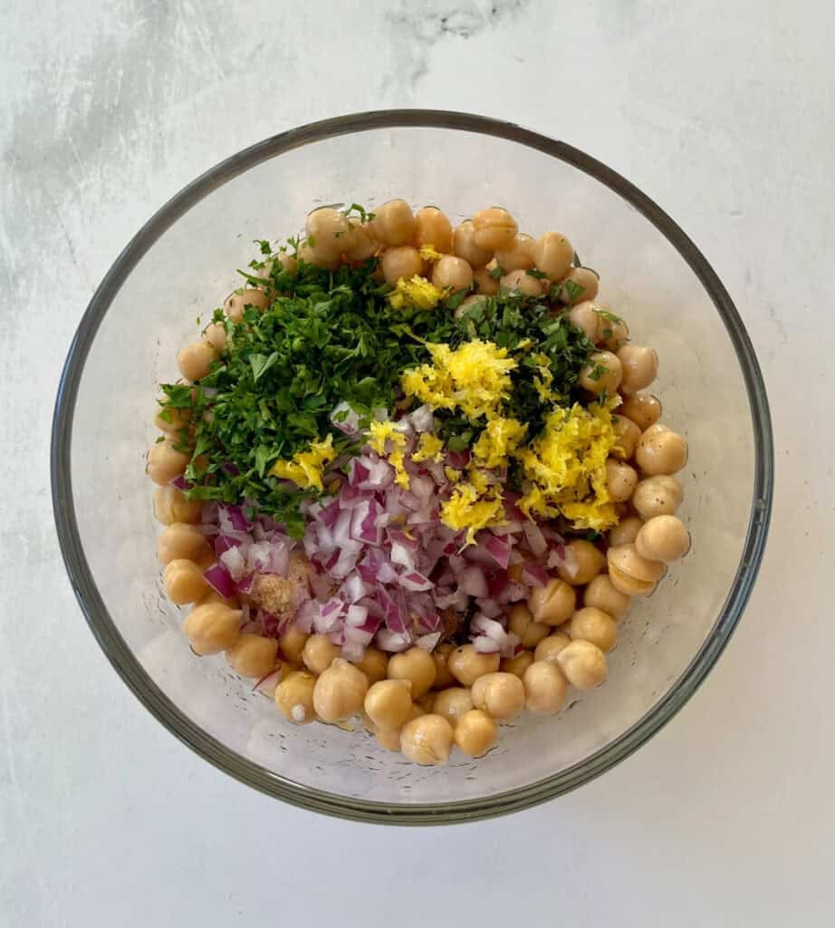 Marinated chickpeas ingredients in a glass bowl with herbs and lemon zest.