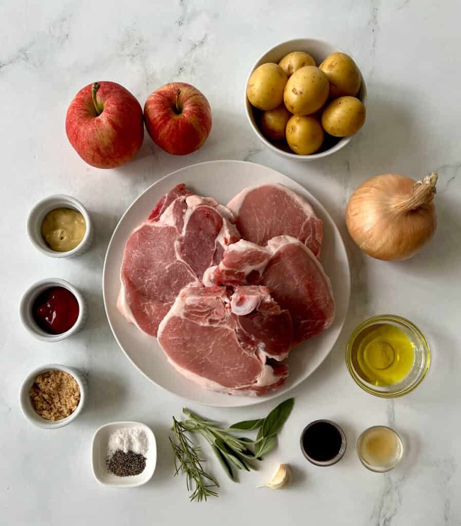 Raw pork chops with apples, potatoes, onion, and herbs on a white surface.
