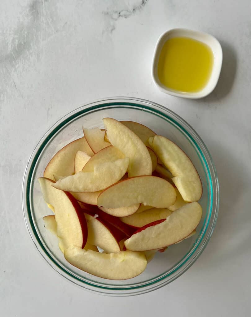 Bowl of sliced apples with a small dish of oil on a white surface.