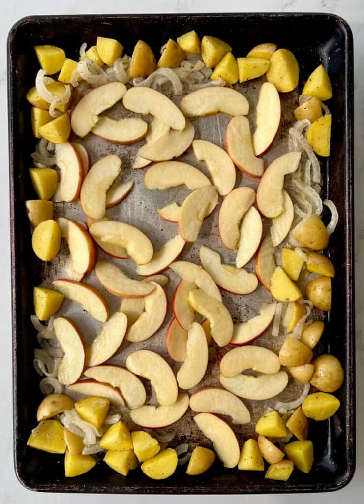 Uncooked roasted vegetables including potatoes, onions, and apples on a baking sheet.