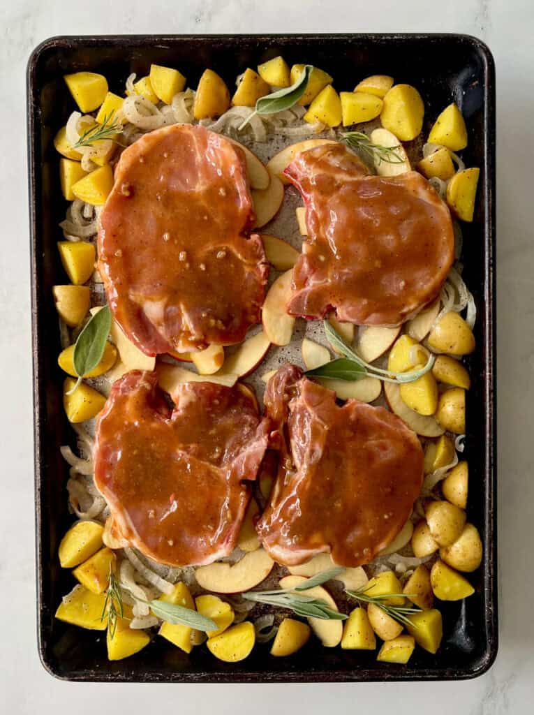 Raw pork chops topped with a sauce with potatoes, apples, onions, and herbs on a baking sheet.