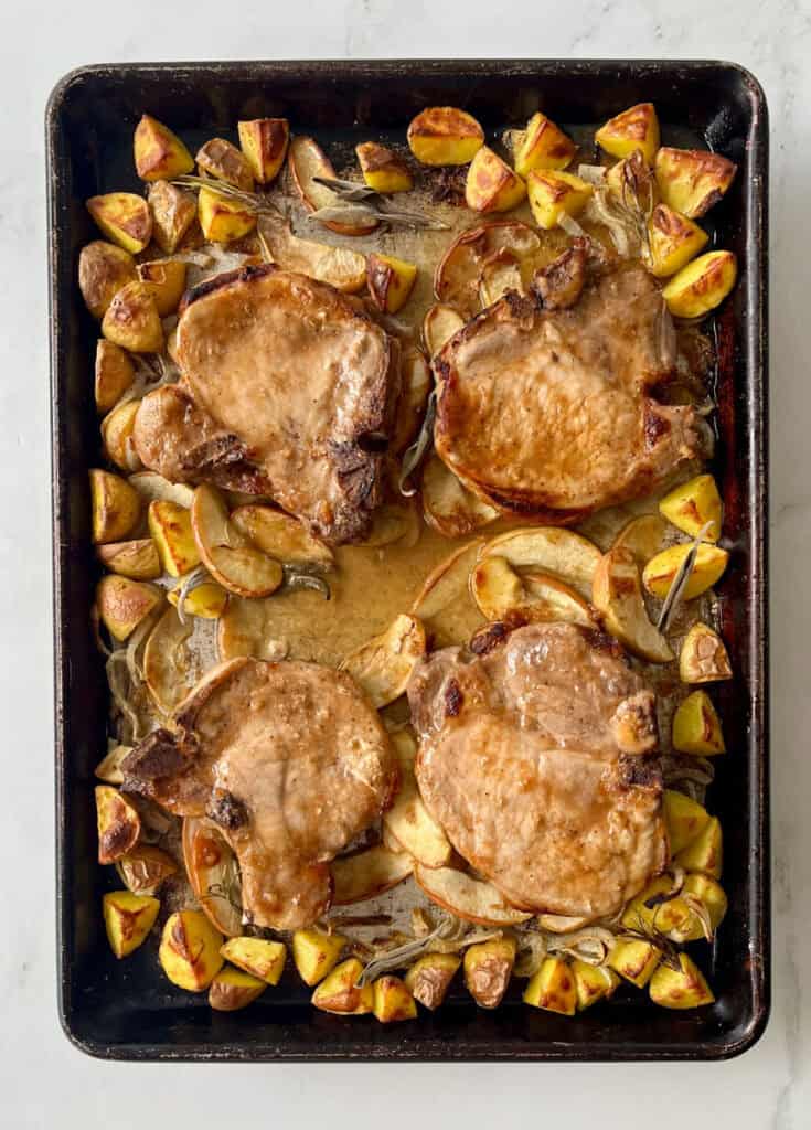 Oven-baked pork chops with roasted potatoes, apples, and onions on a baking sheet.