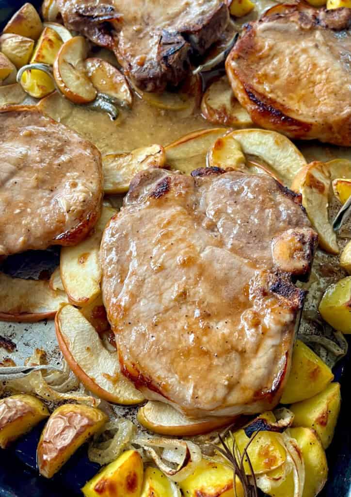 Baked pork chops with roasted potatoes, apples, onions, and herbs on a baking sheet.