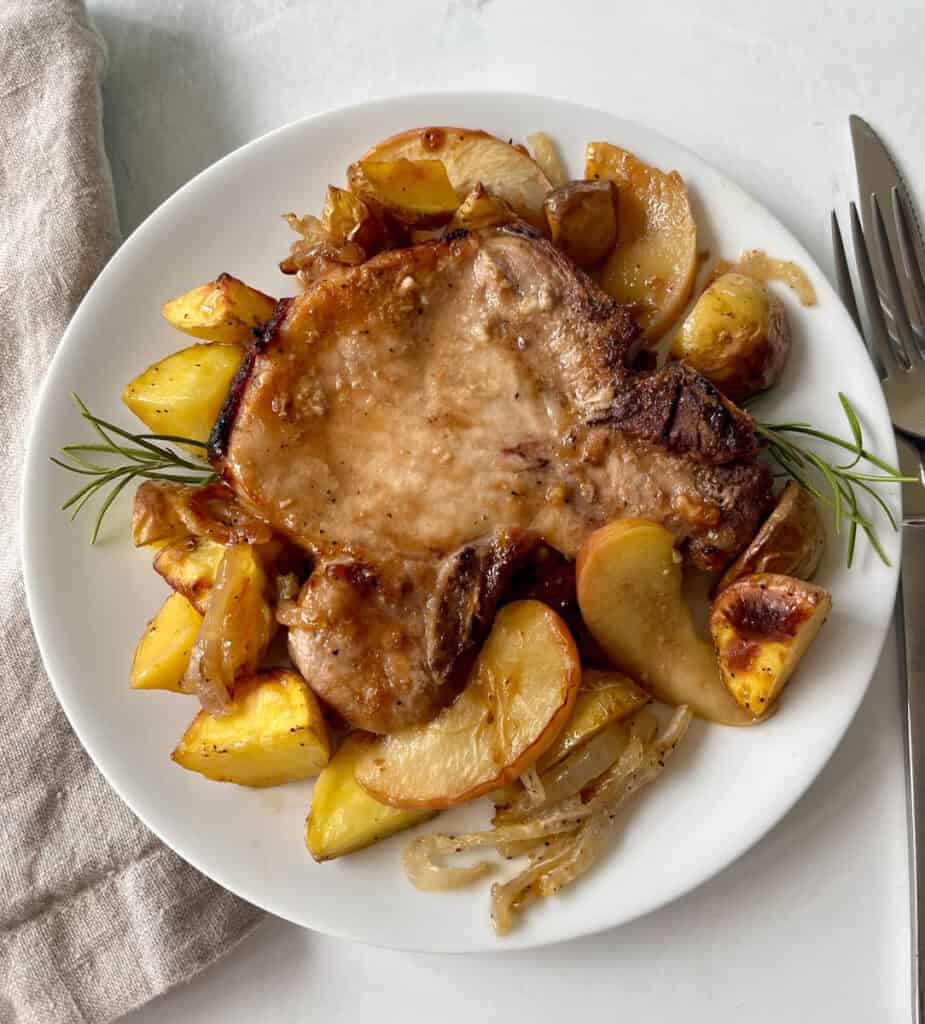 Juicy roasted pork chop served with roasted potatoes, apples, and caramelized onions on a white plate.